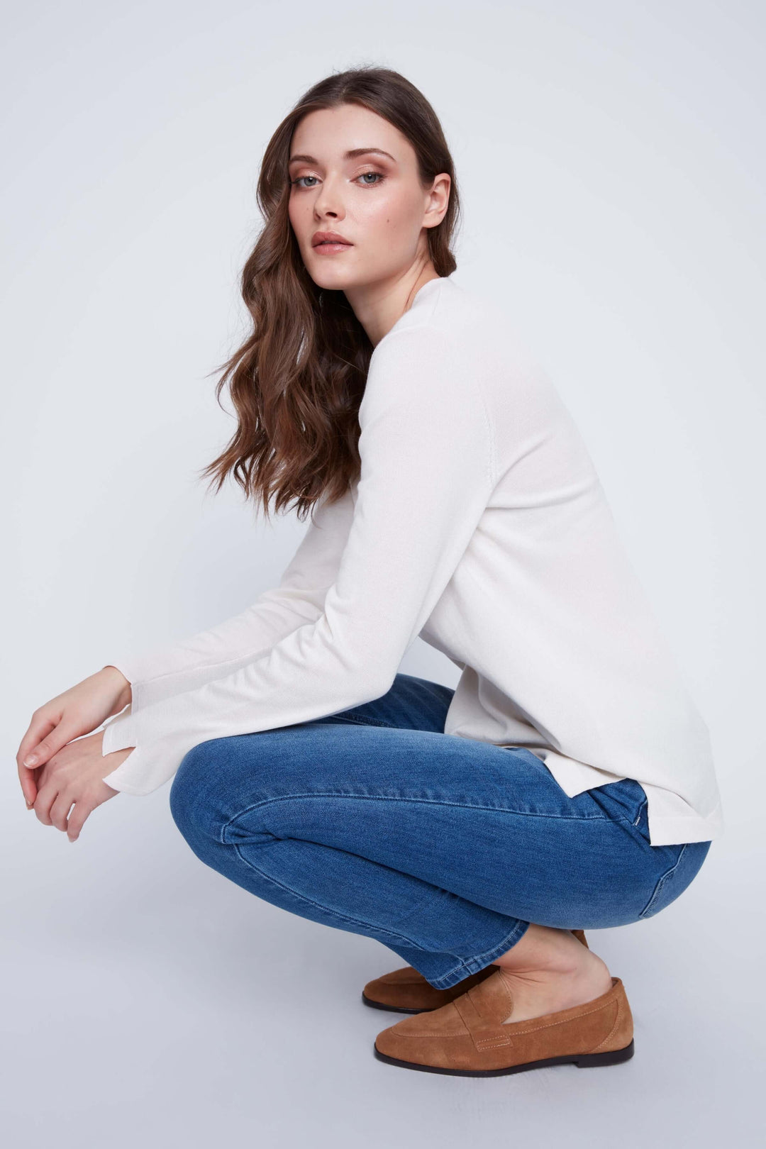 Femme portant un chandail col V blanc avec jeans bleu, assise en squat sur fond neutre.