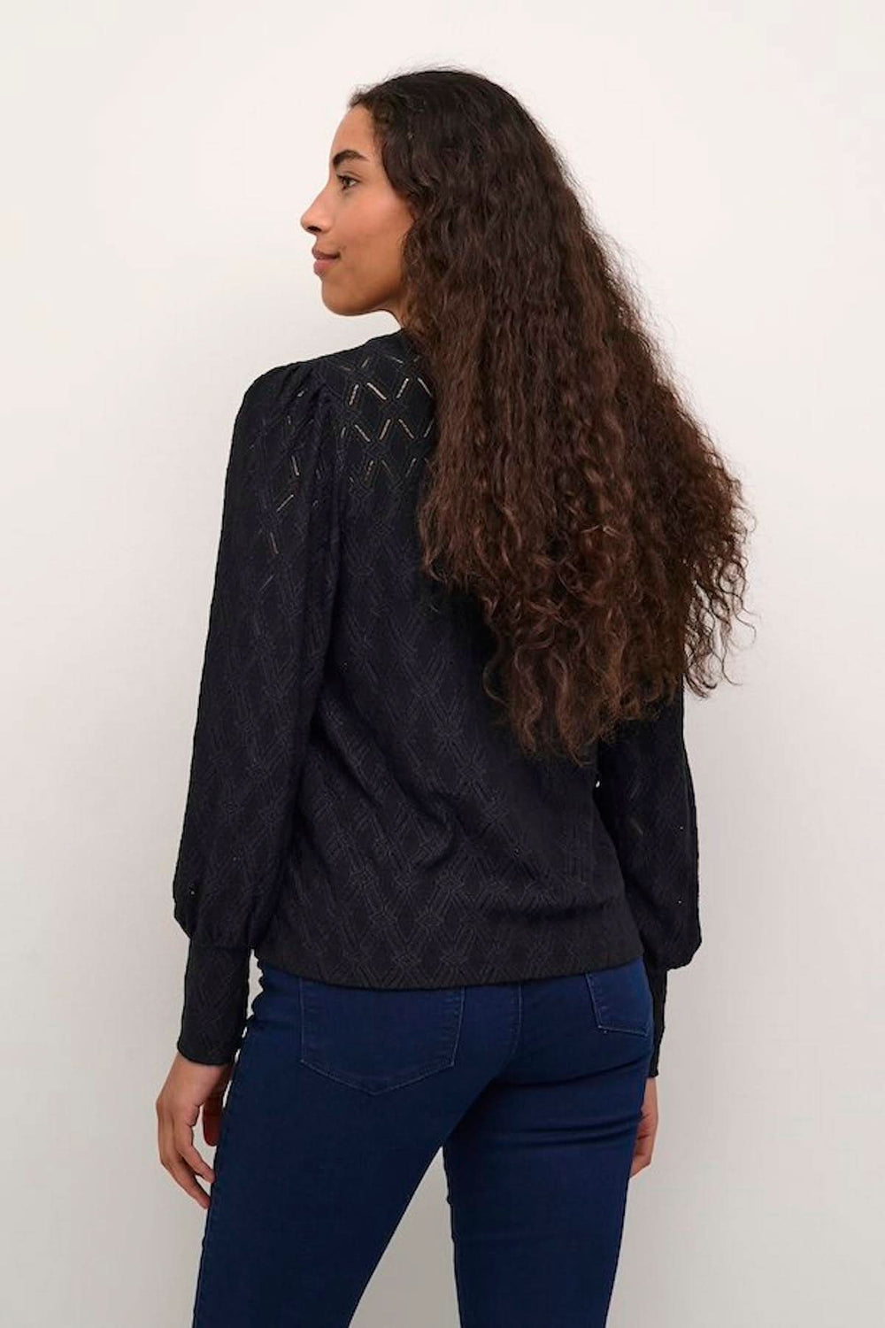 Femme portant une blouse noire texturée avec des manches bouffantes et motifs ajourés, vue de dos.