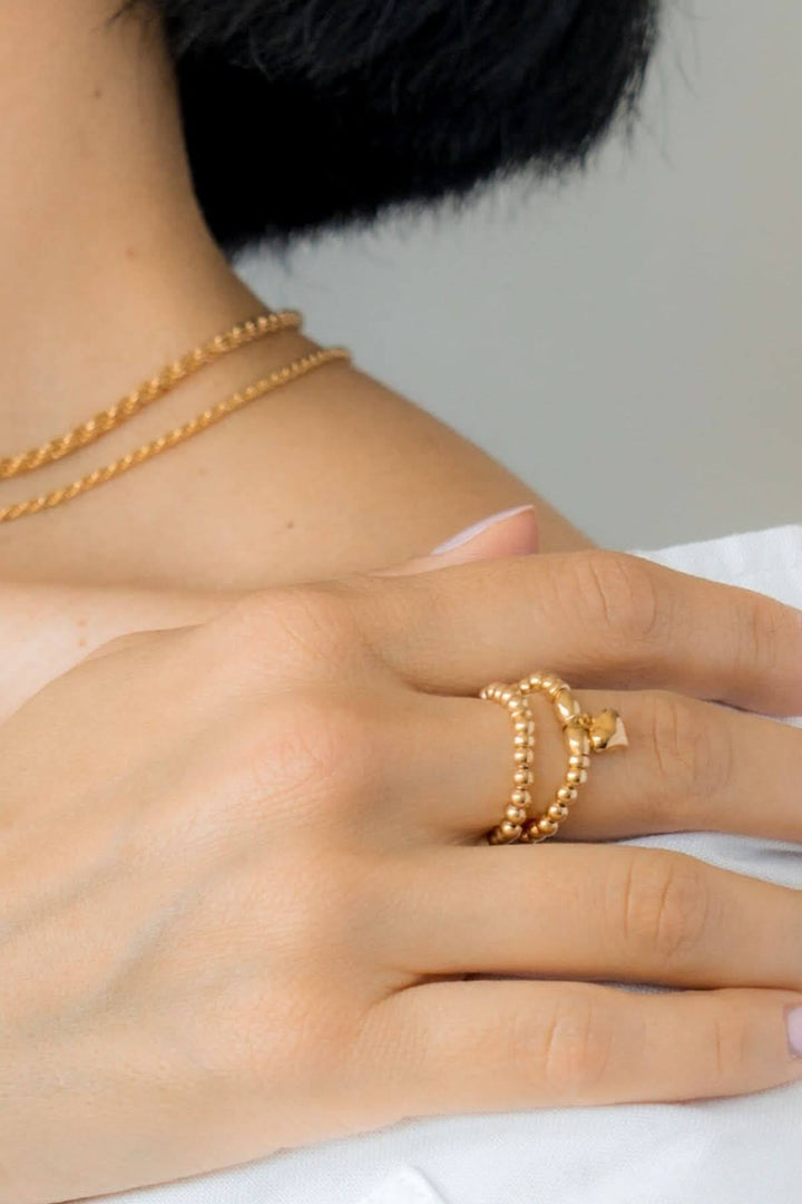 Main d'une femme avec une bague élastique Be Timeless Or et des colliers dorés sur une peau claire.