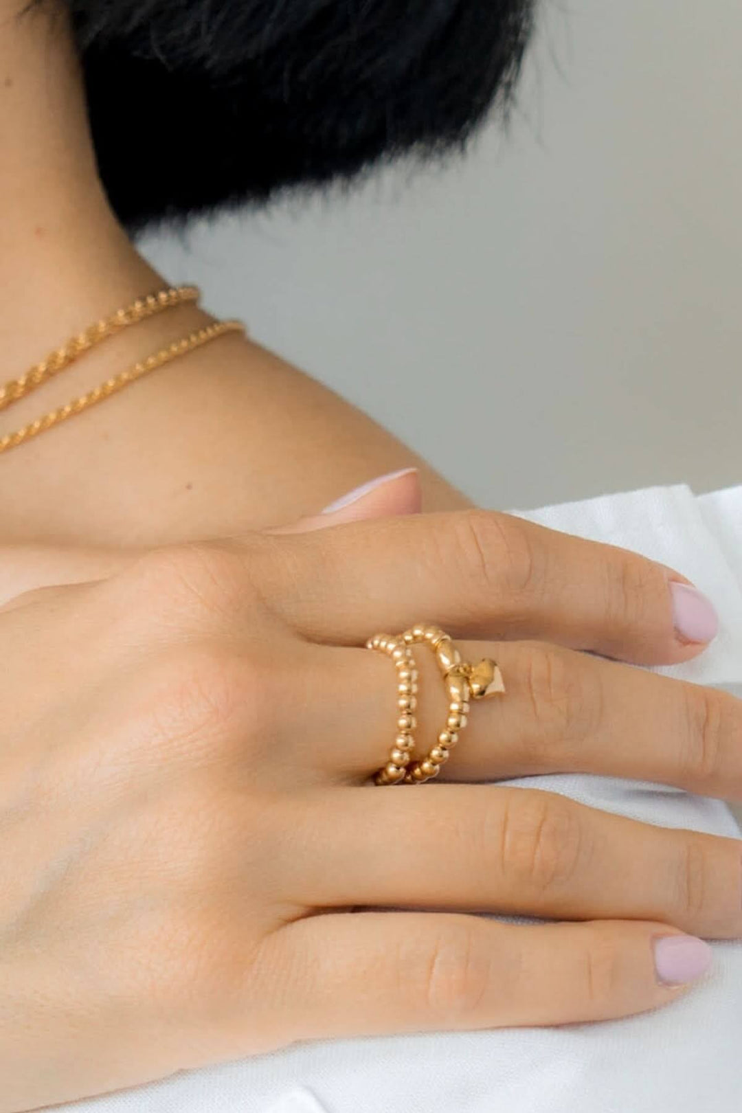 Bague élastique en or Be Tender avec breloque cœur portée sur une main, accompagnée de colliers dorés.