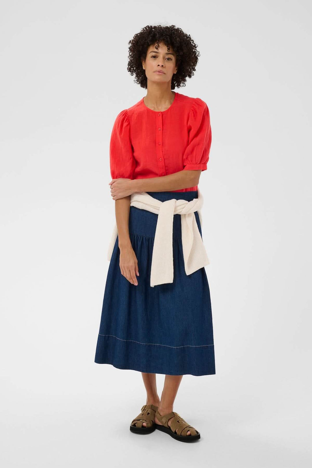 Femme portant Blouse Azra rouge avec manches bouffantes, jupe en denim, et pull autour de la taille, posant debout.