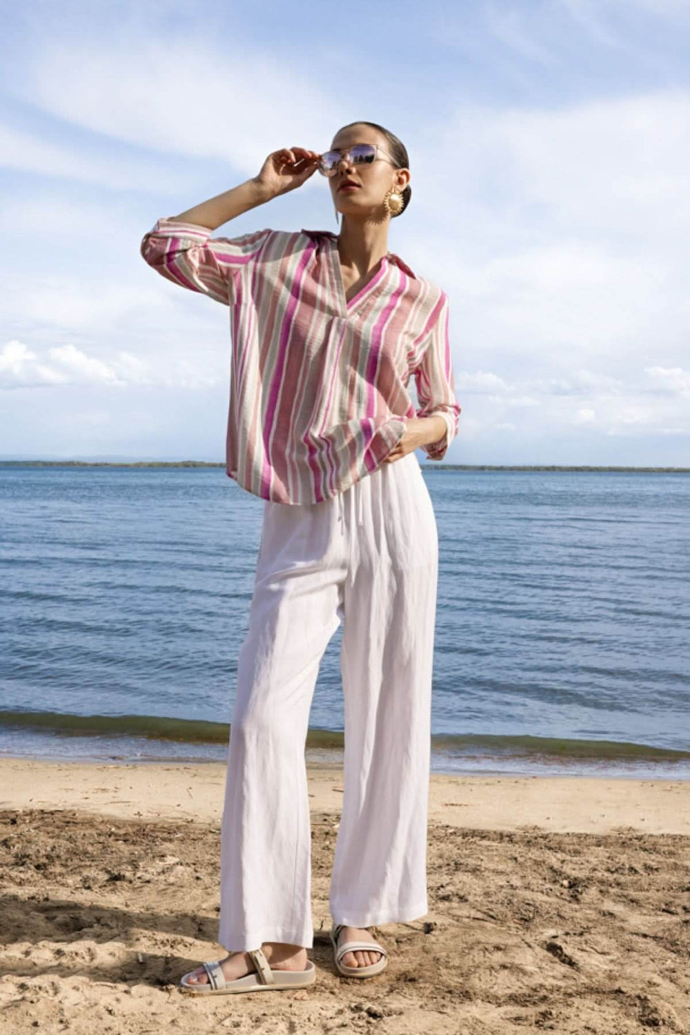 Femme portant une blouse rayée à encolure V sur une plage, illustrant le style élégant et décontracté avec manches trois-quarts.
