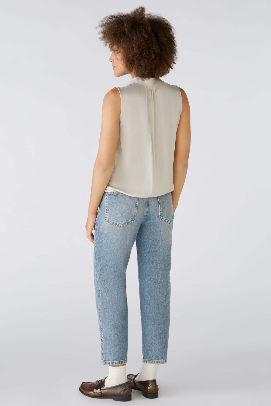 Person wearing a sleeveless cream blouse with light blue jeans and brown loafers, viewed from the back.