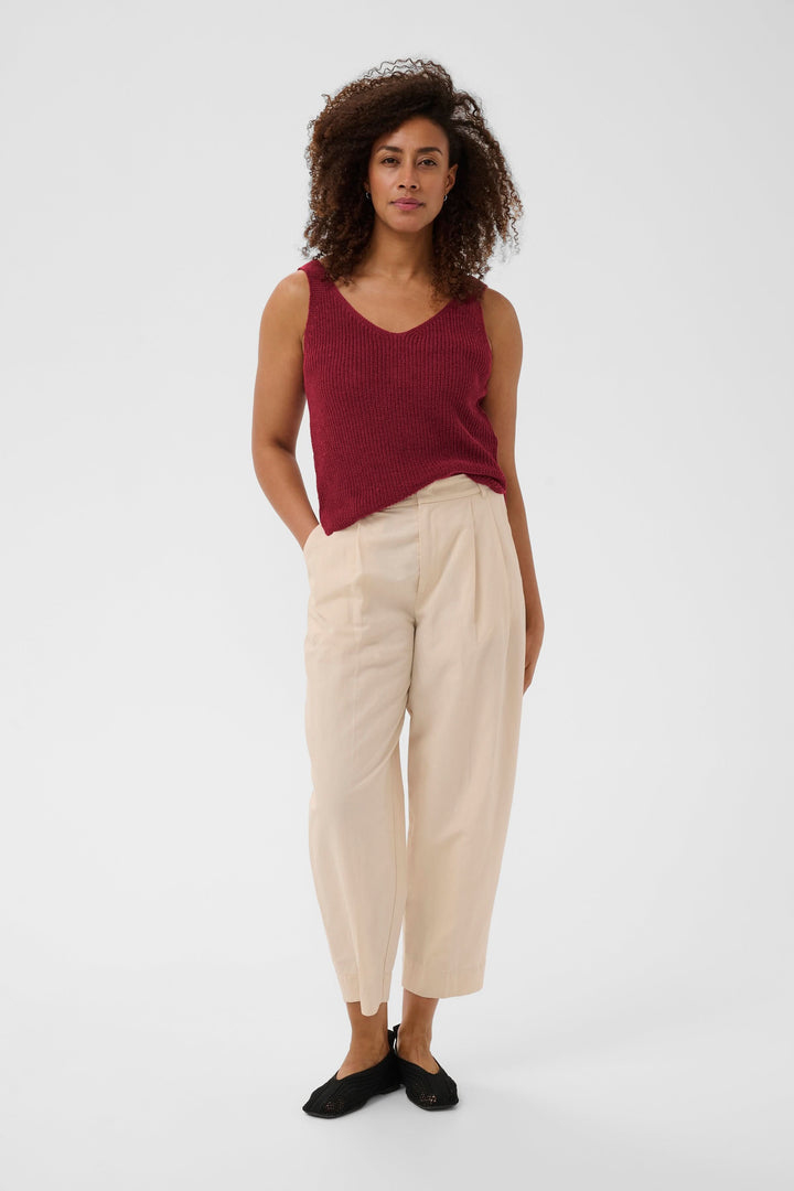 Femme portant une camisole Rebecca tricotée col v sans manches rouge, style décontracté, avec un pantalon beige et des chaussures noires.