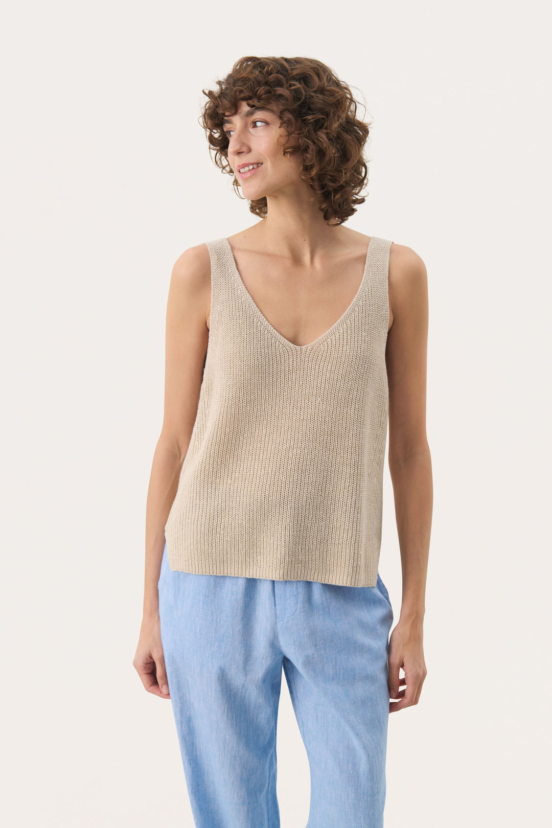 Femme portant une camisole Rebecca tricotée col v sans manches en maille légère, couleur beige, idéale pour une tenue estivale décontractée.
