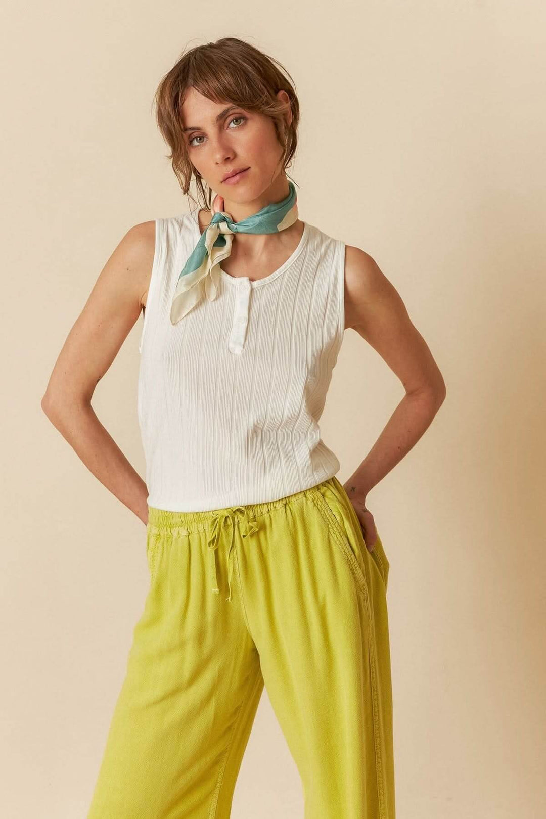Femme portant un débardeur en maille côtelée blanche avec une écharpe, pantalon jaune, fond neutre.