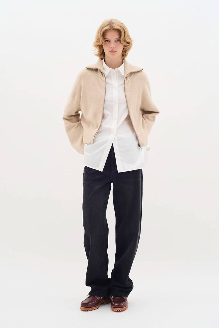 Cardigan Jogan beige à fermeture zippée, col montant, porté avec une chemise blanche et un pantalon noir.