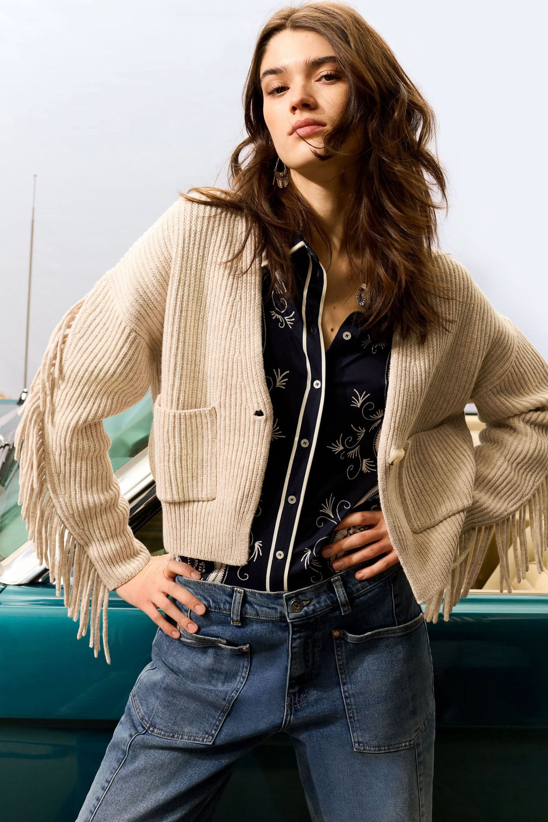 Cardigan avec franges beige, manches amples, décolleté en V, porté avec une chemise à motifs, style moderne.