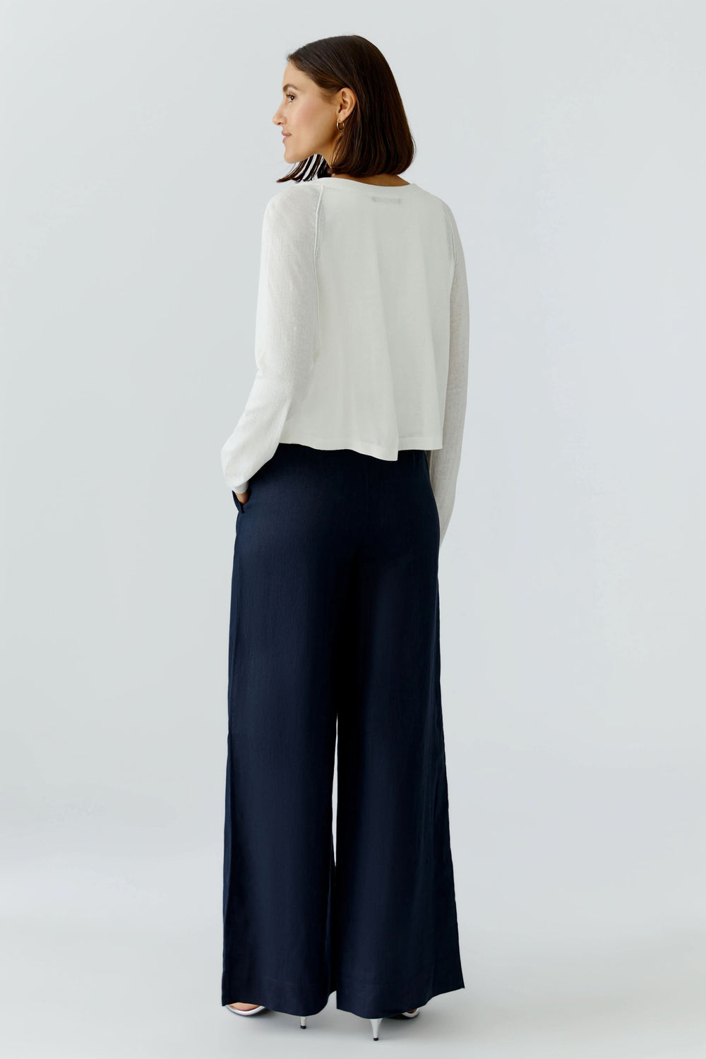 Femme portant un cardigan texturé boutonné blanc avec un pantalon large bleu foncé, vue de dos en studio.