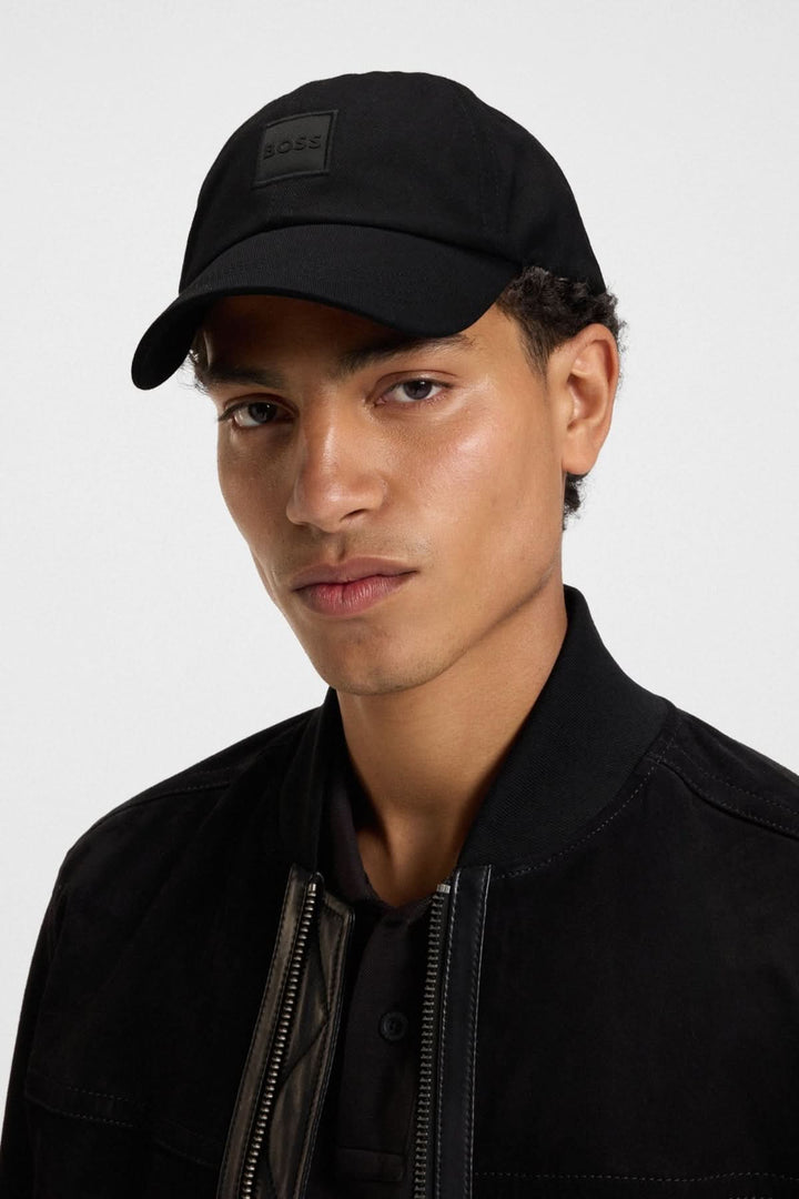 Homme portant une casquette BOSS noire avec écusson ton sur ton, style contemporain, vêtement décontracté.