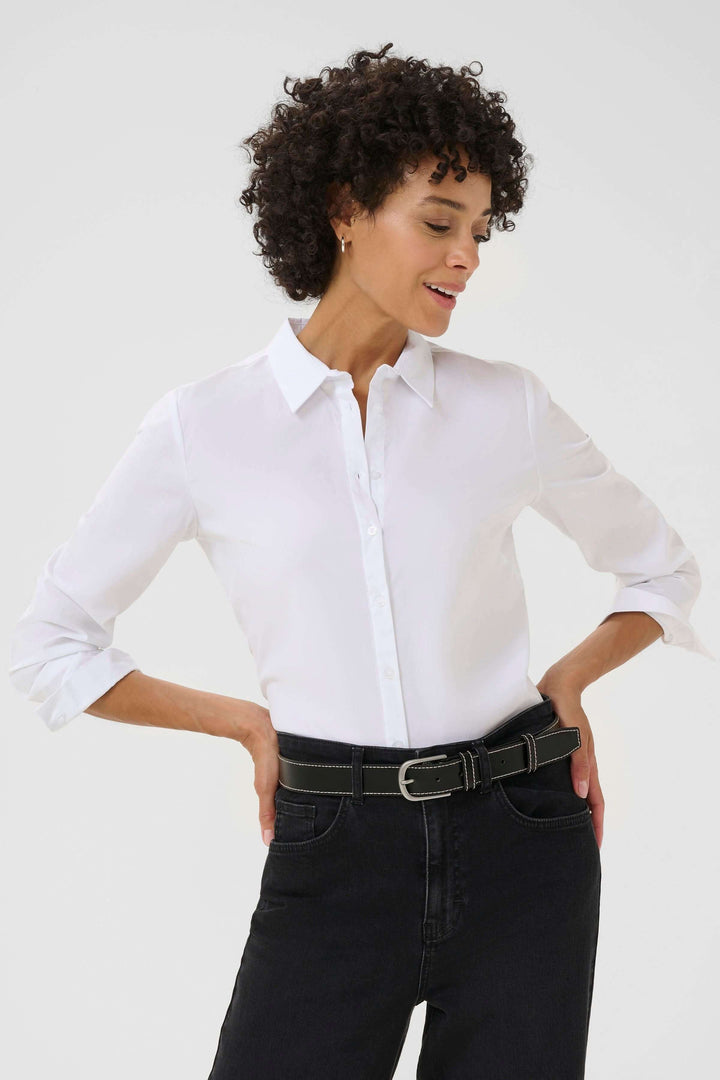 Femme souriante portant une chemise blanche avec une ceinture en cuir noire et un pantalon noir.