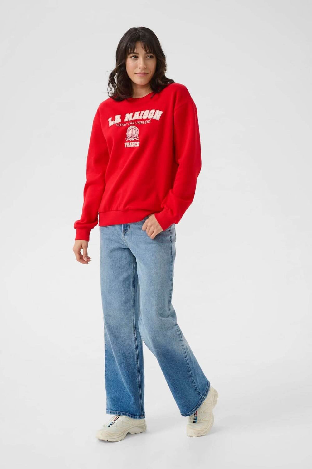 Chandail à col rond rouge avec inscription brodée, porté avec un jean décontracté et des chaussures blanches.