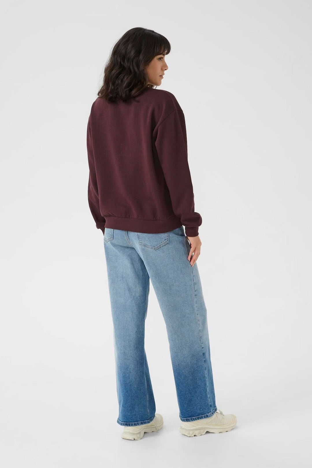 Femme portant un chandail à col rond bordeaux avec des jeans, vue de dos sur fond clair.