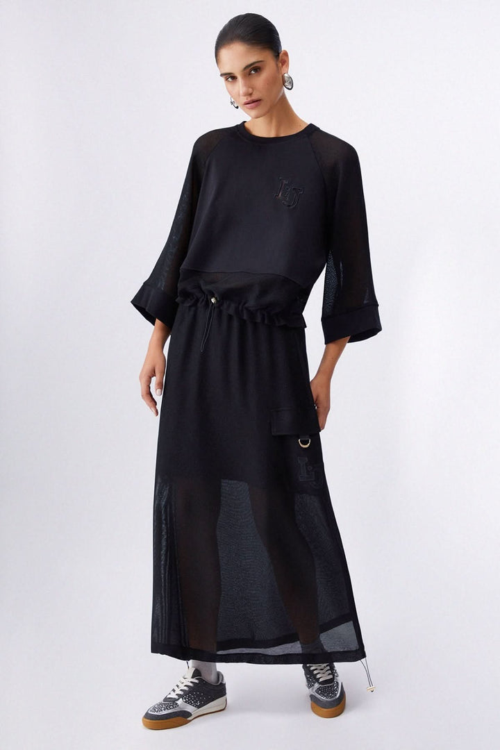 Femme portant un chandail court à manches larges noir avec une jupe longue, style moderne et décontracté, détail écusson logo.