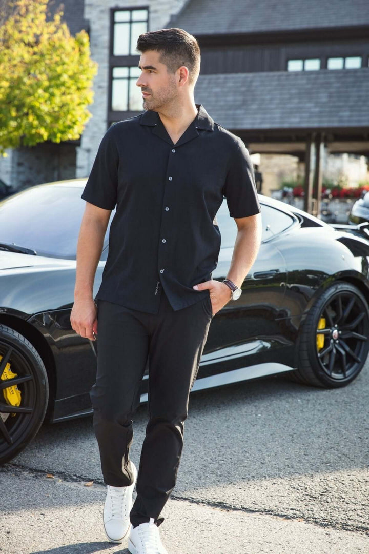 Homme portant une chemise noire à col cubain et pantalon noir, debout près d'une voiture sportive.