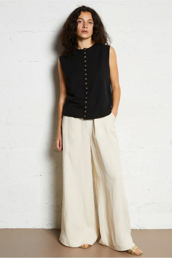 Femme portant une chemise noire sans manches et un pantalon blanc ample, posant contre un mur blanc.