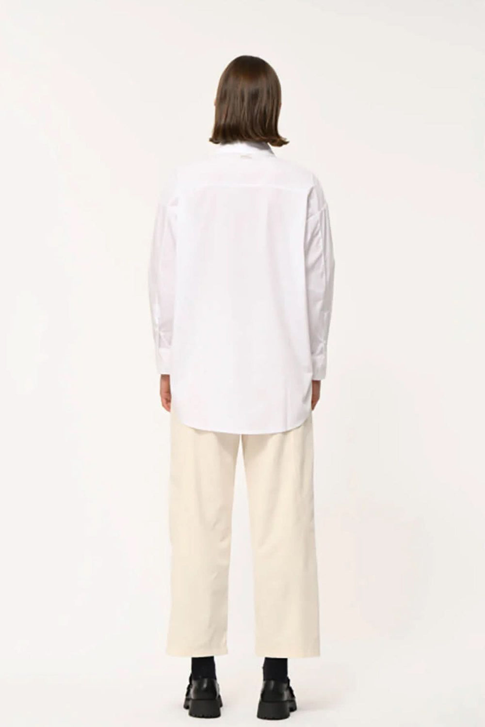 Dos d'une chemise classique à coupe droite, ample, col pointu dissimulé, manches longues, portée sur un pantalon clair.