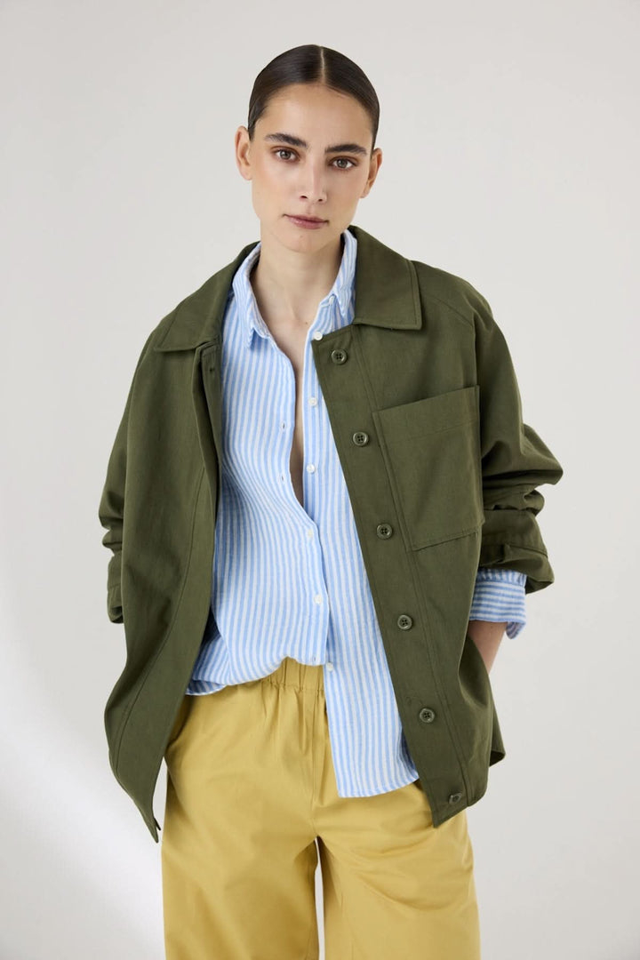 Chemise rayée à col arrondi sous une veste kaki, modèle debout, mains dans les poches, style décontracté et moderne.