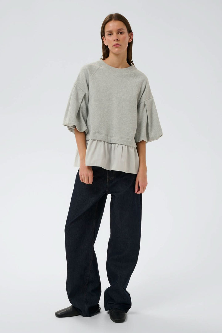 Femme portant un haut ouaté avec ourlet Dessy, coupe ample, manches volumineuses et ourlet plissé, silhouette moderne et confortable.