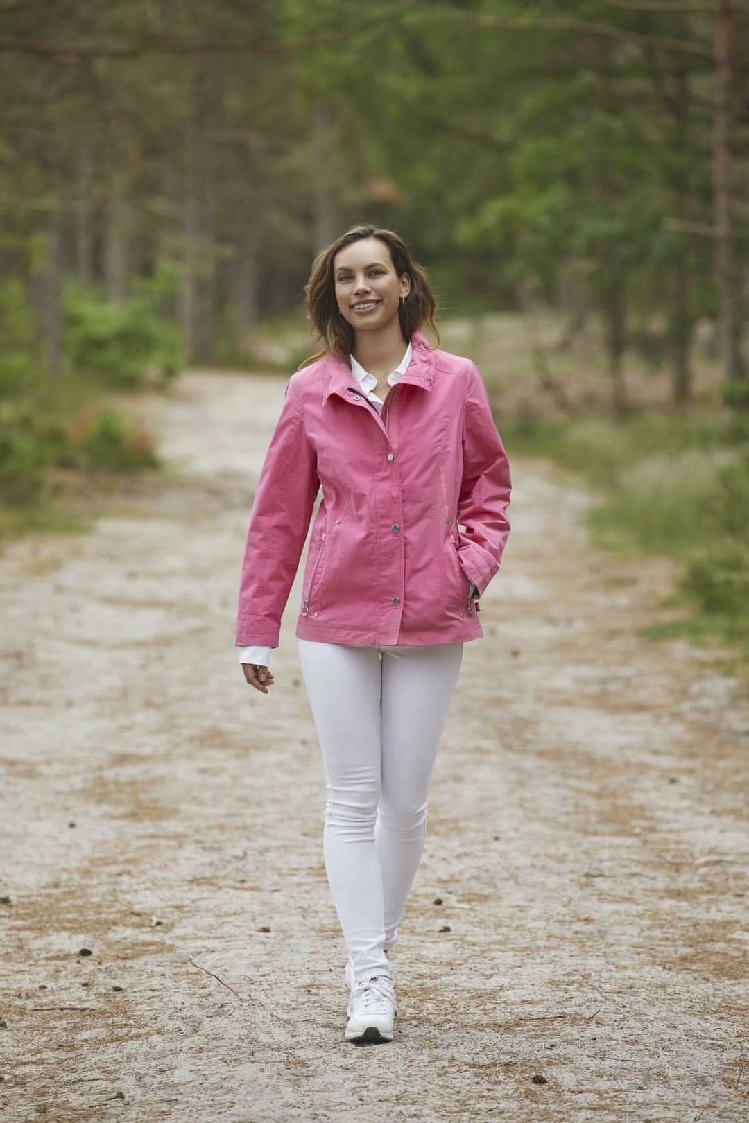 Femme marchant sur un chemin, portant un imperméable sans capuchon rose et un pantalon blanc, en pleine nature.
