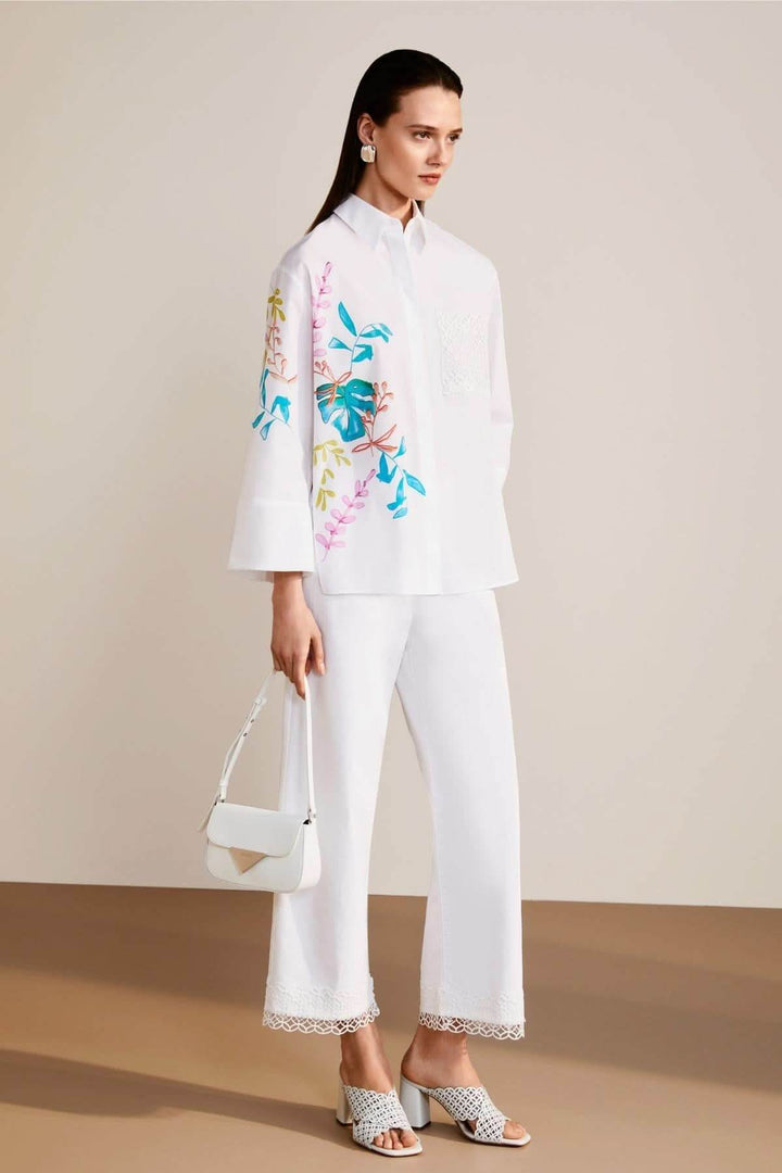 Femme en chemisier blancc avec broderies colorées et pantalon blanc aux détails en dentelle.