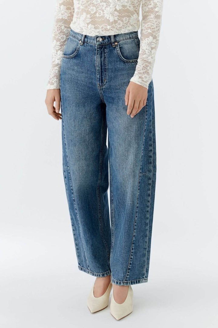 Jean baril taille haute en denim bleu, coupe baril avec jambe arrondie, porté avec un haut en dentelle blanche.