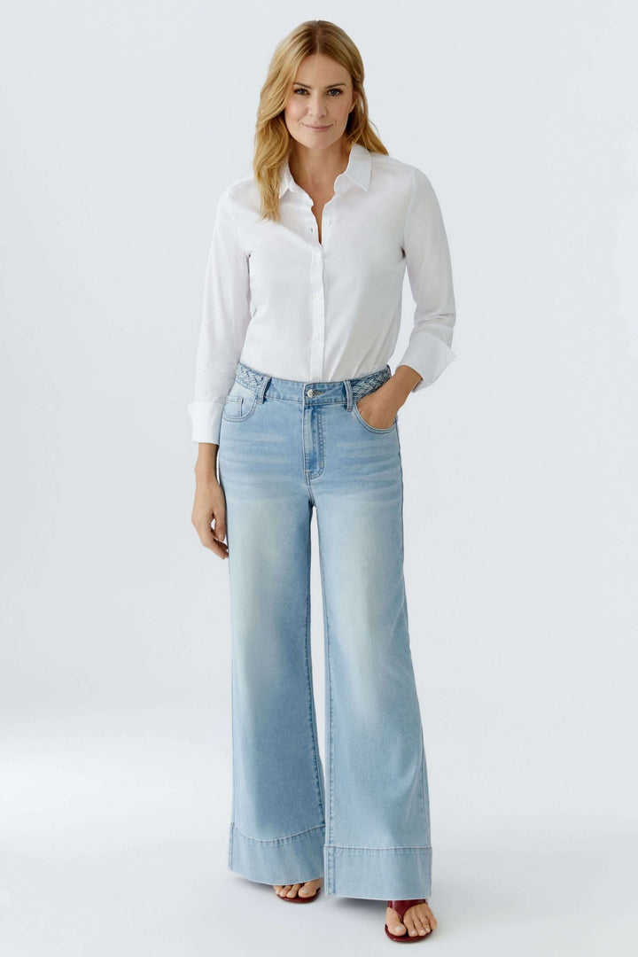 Jean à jambe large avec taille tressée en denim, porté avec une blouse blanche. Silhouette moderne et détail élégant.