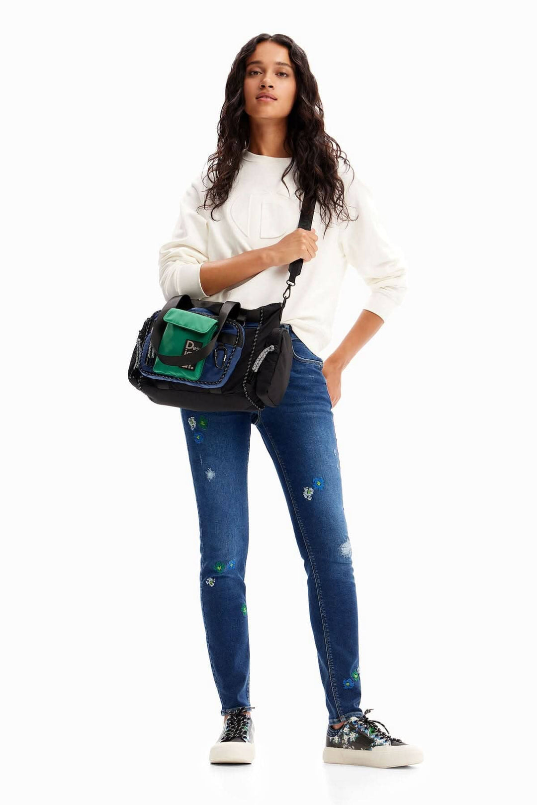 Femme en jean skinny brodé, sweatshirt blanc et sac de mode, pose debout avec style.