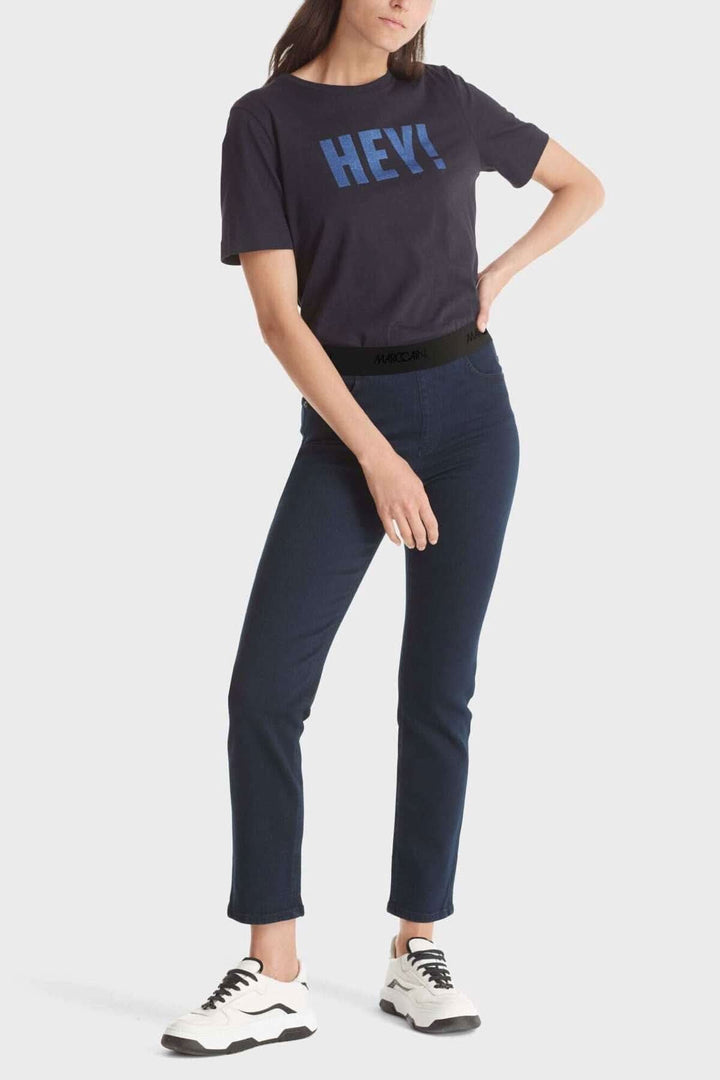 Femme portant un t-shirt noir avec le texte bleu "HEY!" et un pantalon SIENA en denim bleu.