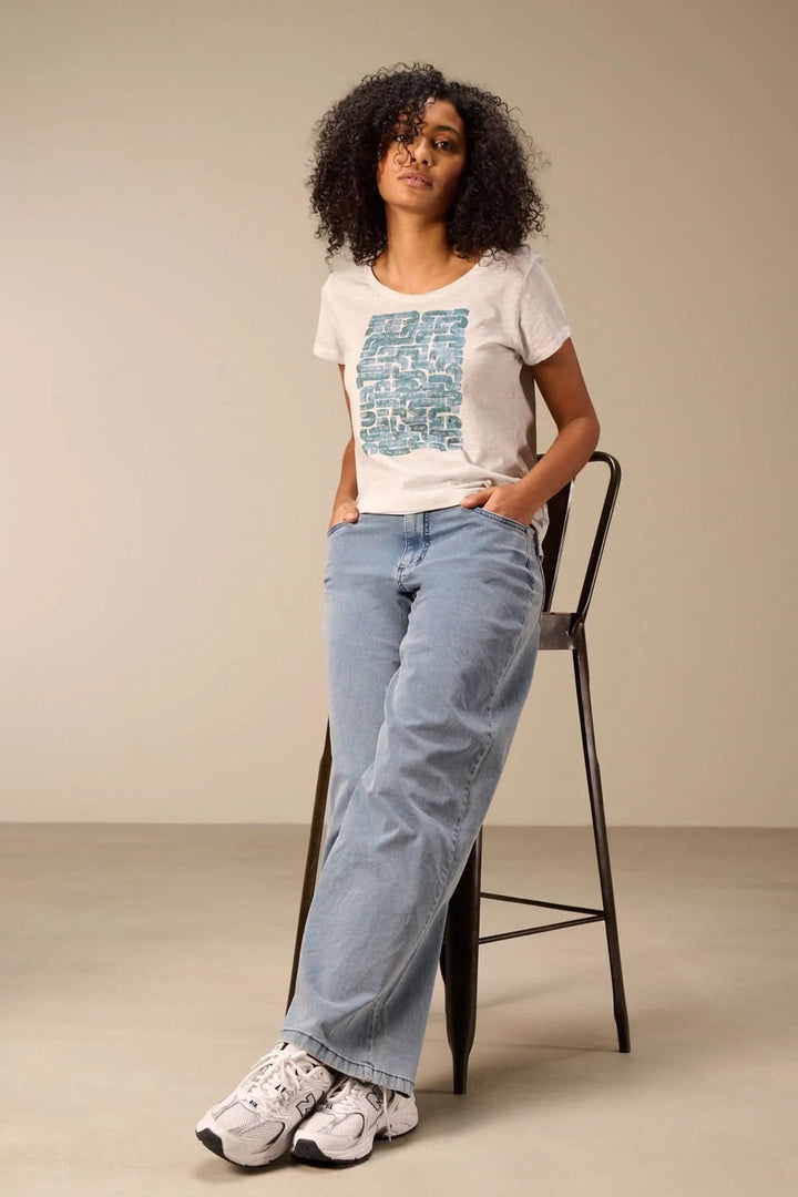 Femme portant un jeans jambe large évasée et un t-shirt décontracté, posée sur un tabouret avec des baskets blanches.