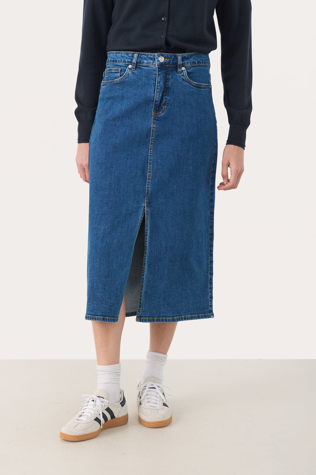 Jupe Dillin en denim bleu, midi avec fente frontale, portée avec un chandail et des baskets.