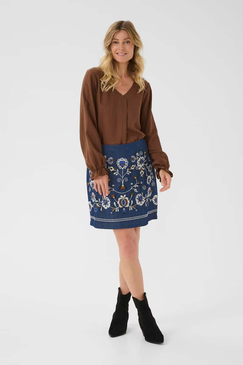 Jupe Gro en denim avec broderies florales, parfait pour un style bohème chic, ici portée avec un haut marron et des bottines noires.