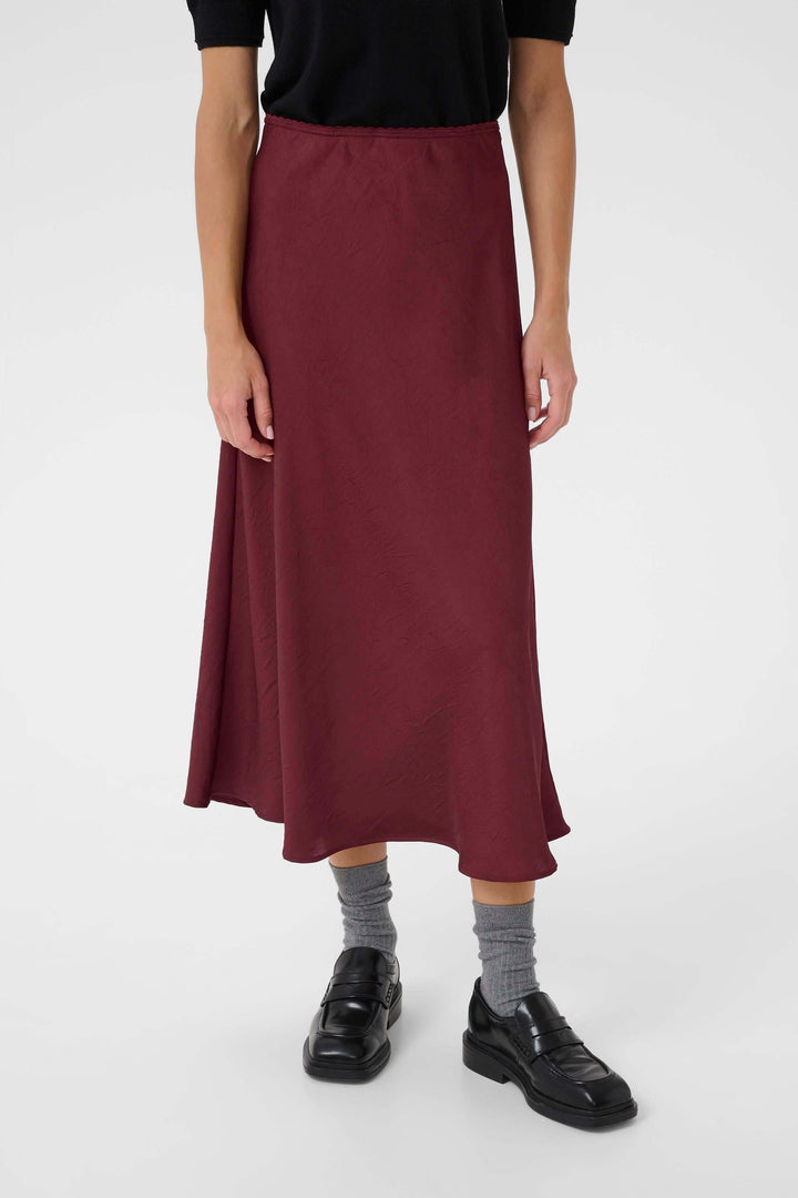 Jupe midi fluide rouge bordeaux avec coupe évasée, portée avec un haut noir et des chaussures noires.