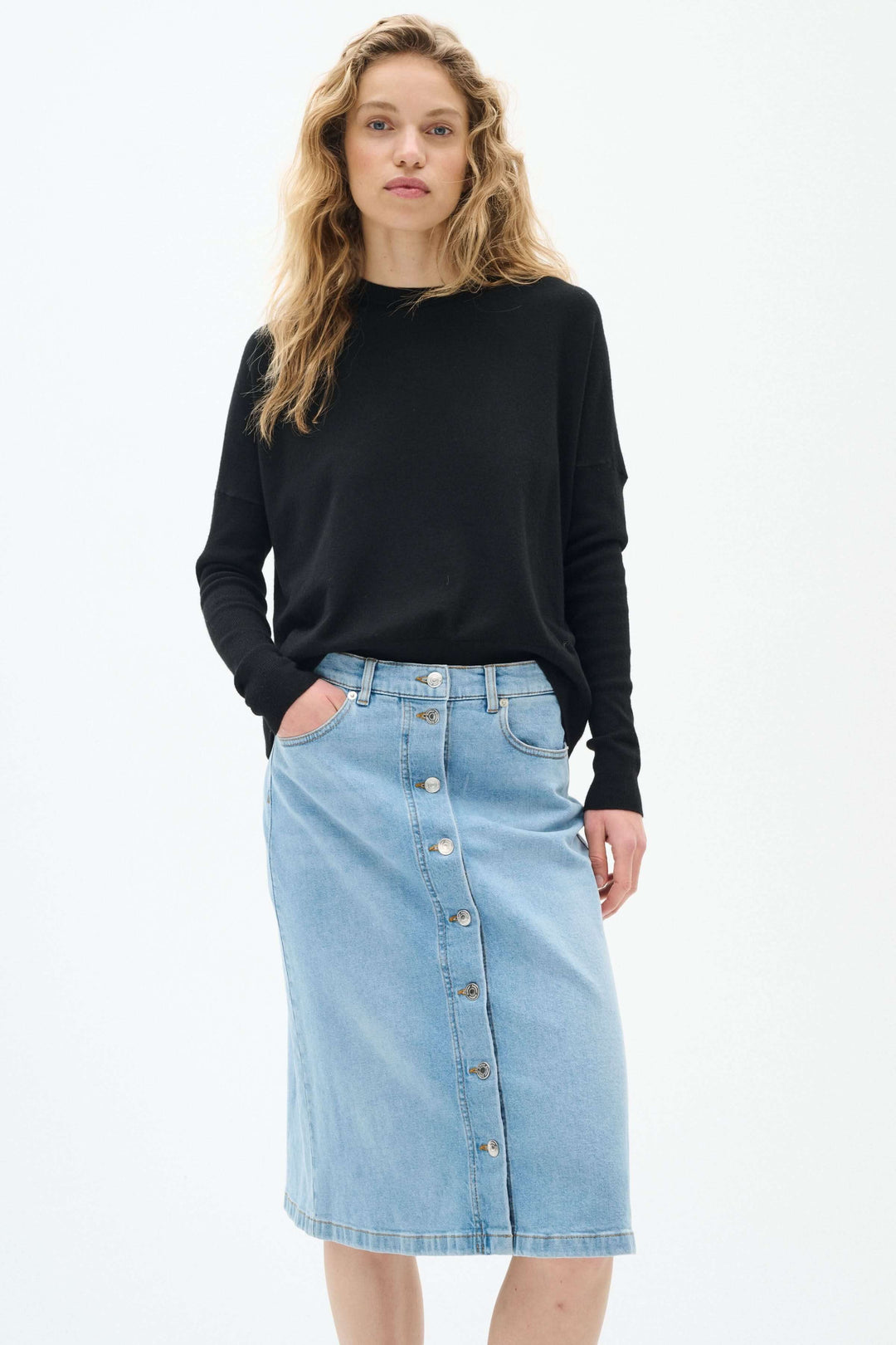 Jupe en jeans bleu clair à boutons, coupe droite, longueur midi, portée avec un pull noir.