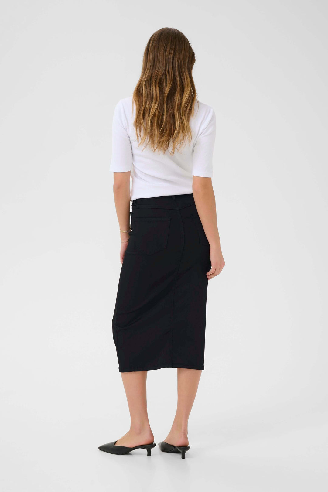Femme portant une jupe longue Dima en denim noir, vue de dos, avec un t-shirt blanc et des mules noires.