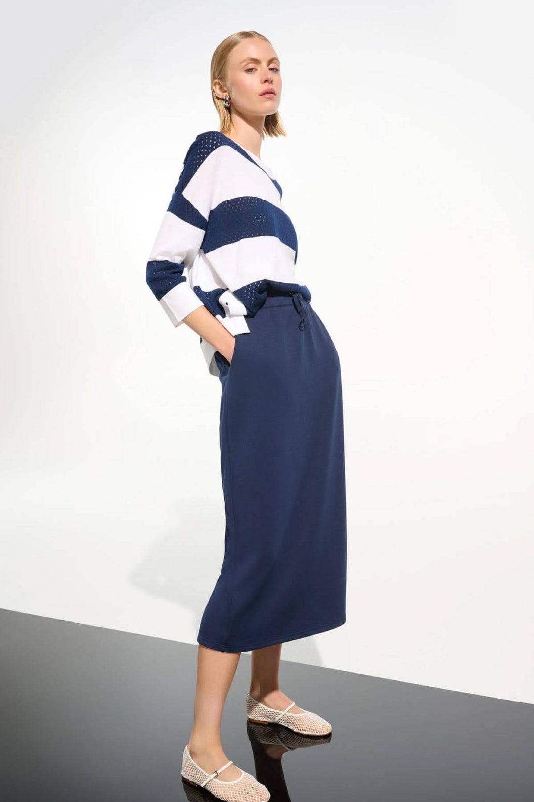 Femme portant une jupe midi à taille coulissante et un chandail rayé, style décontracté et moderne, longueur mi-mollet, chaussures plates.