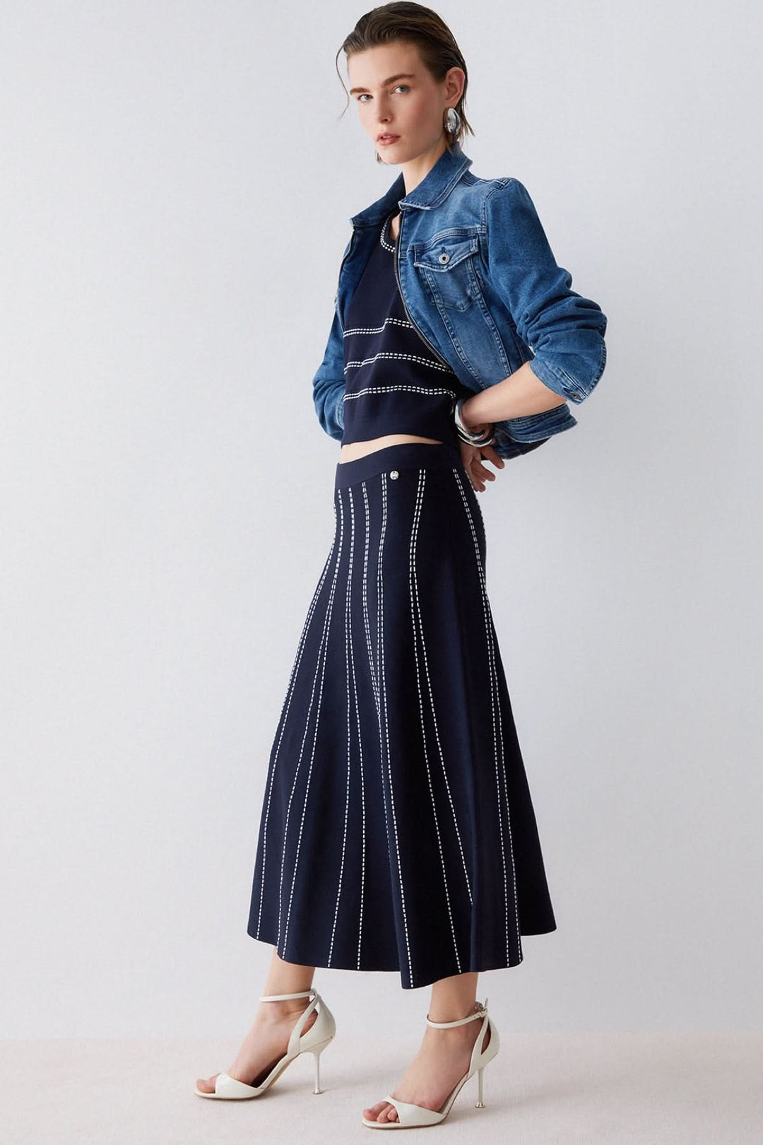 Femme portant une jupe midi évasée à motif rayé, associée à une veste en jean et des talons hauts, look élégant et moderne.
