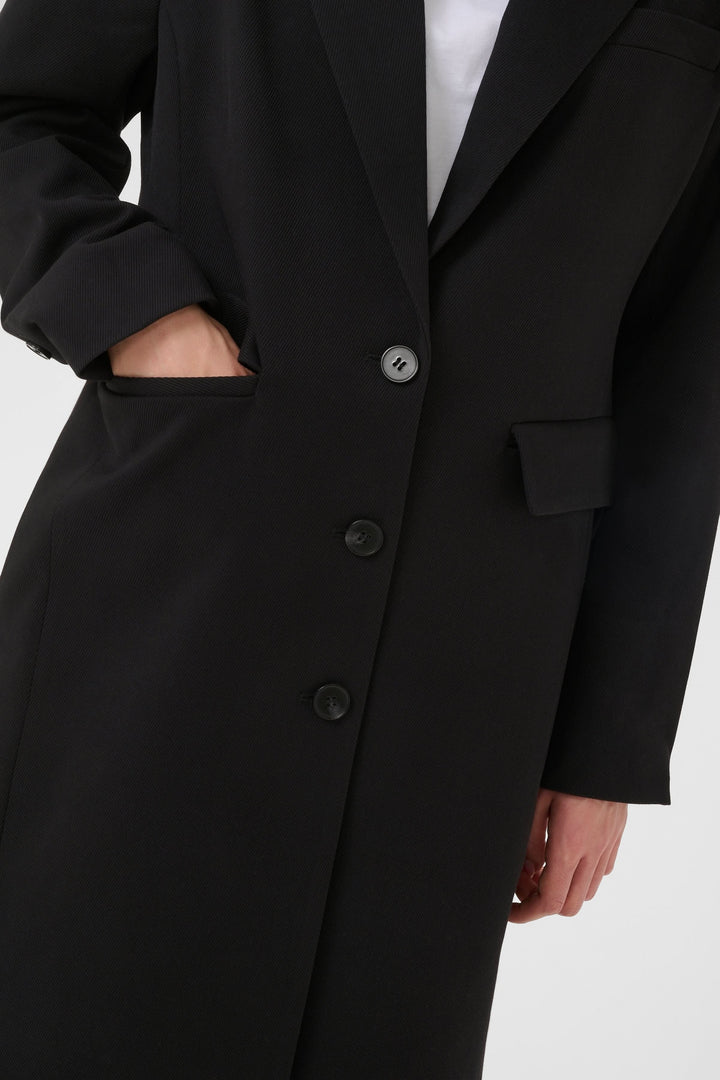 Manteau Milos noir, silhouette droite et structurée, long, raffiné, col tailleur, boutons, tenue élégante et intemporelle.