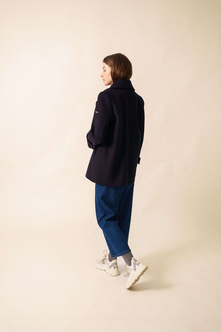 Femme portant le manteau chic STE ROMY, longueur sous les fesses avec col en V, deux poches et boutons, sur un fond neutre.