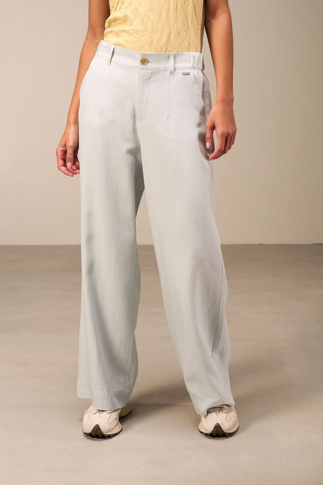 Pantalon large fluide bleu clair avec taille haute, plis marqués et fermeture à bouton, porté avec un haut jaune.