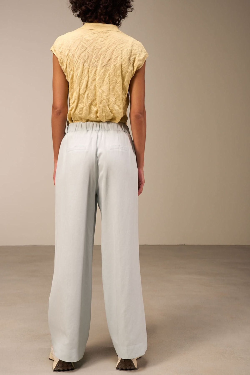 Femme vue de dos portant un pantalon large bleu clair et un haut jaune, dans un intérieur minimaliste.