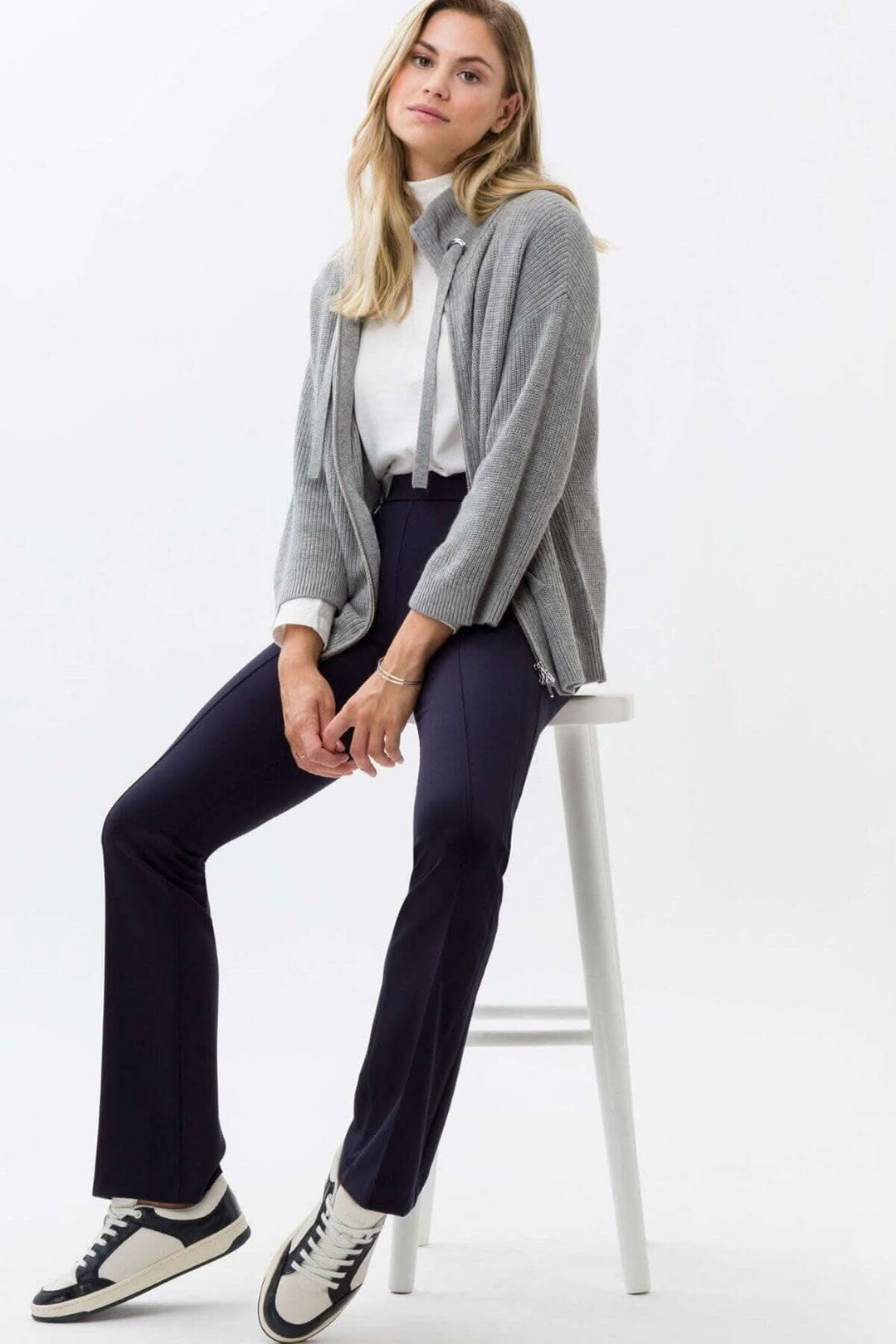 Femme assise sur une chaise, portant un cardigan gris et des pantalons skinny bootcut, complet avec des baskets blanches.