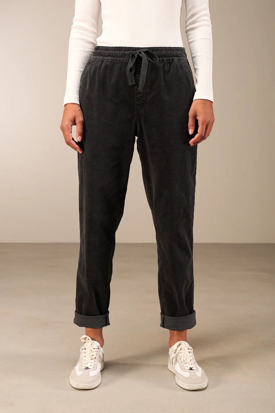 Pantalon en velours côtelé noir avec taille élastique, jambe fuselée, retroussé, porté avec des baskets blanches et un haut blanc.