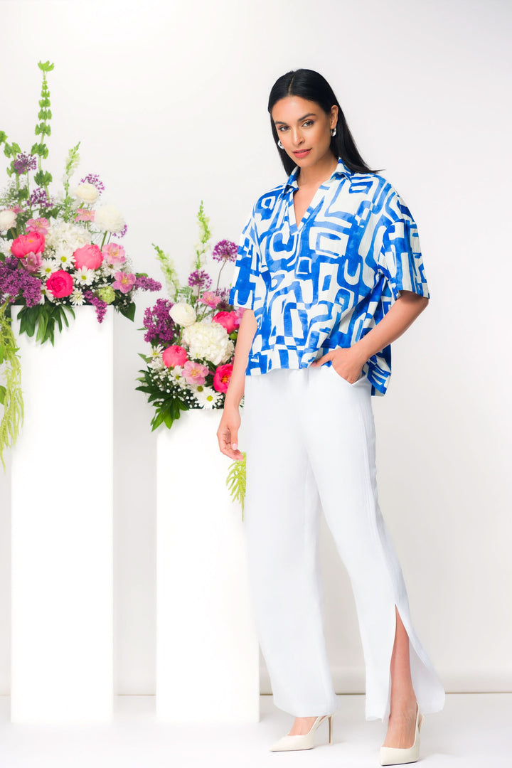 Femme portant un pantalon fluide à fente latérale et une chemise bleue à imprimé géométrique, posant devant des fleurs colorées.