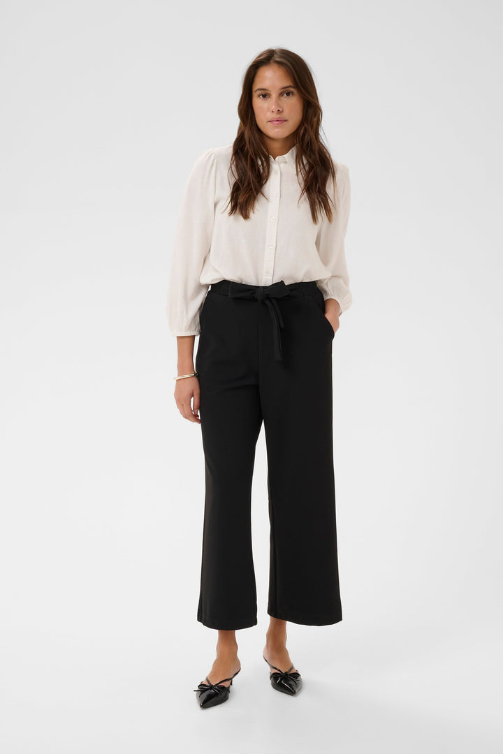 Femme portant un pantalon jambe large Phoebe avec ceinture intégrée, blouse blanche élégante et chaussures noires, look raffiné et polyvalent.