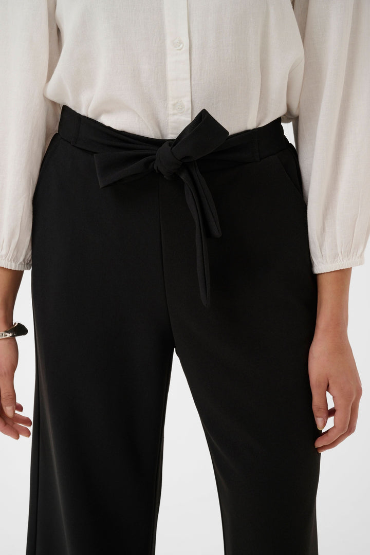 Pantalon jambe large Phoebe noir avec ceinture intégrée nouée, porté avec une blouse blanche.
