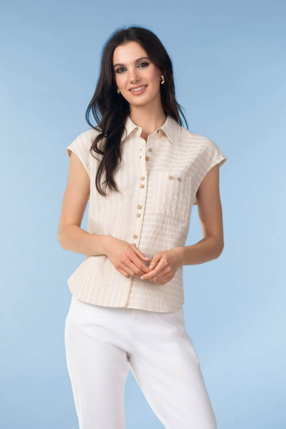 Femme portant un chemisier beige et un pantalon blanc, posant sur un fond bleu clair.