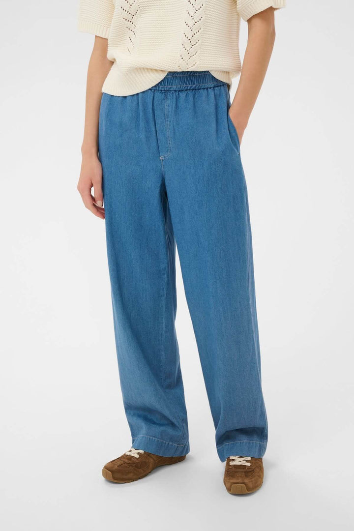 Pantalon taille élastique Aina en denim léger avec coupe droite et taille élastique, porté avec un haut crème et des chaussures marron.