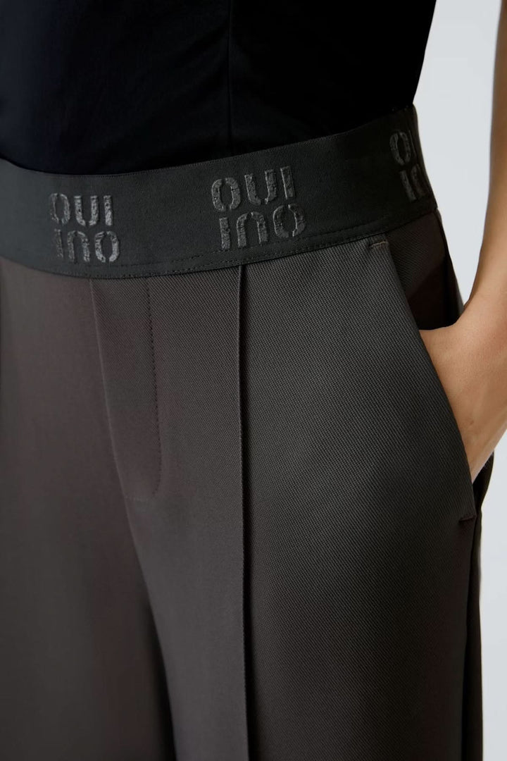 Pantalon à taille élastique avec coupe ample, plis marqués à l'avant, couleur foncée et ceinture noire, confort et élégance moderne