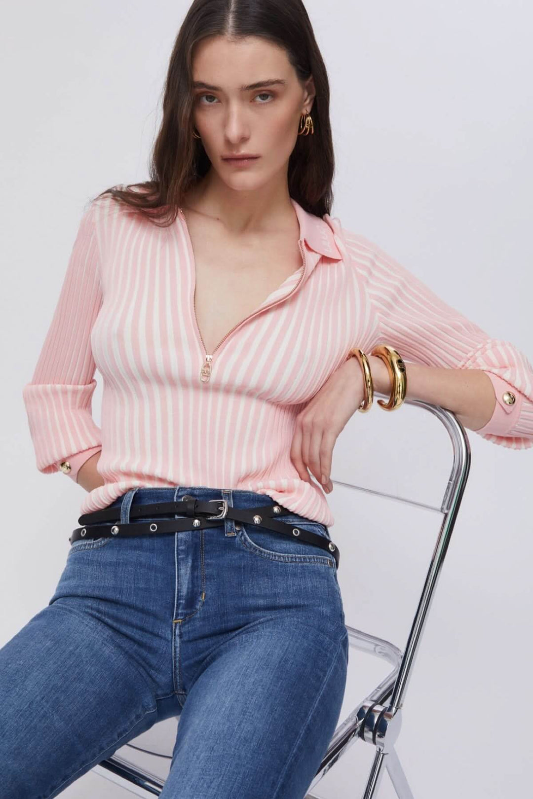 Femme portant un polo en maille côtelée à demi-glissière à rayures roses, assise sur une chaise, avec des accessoires dorés.